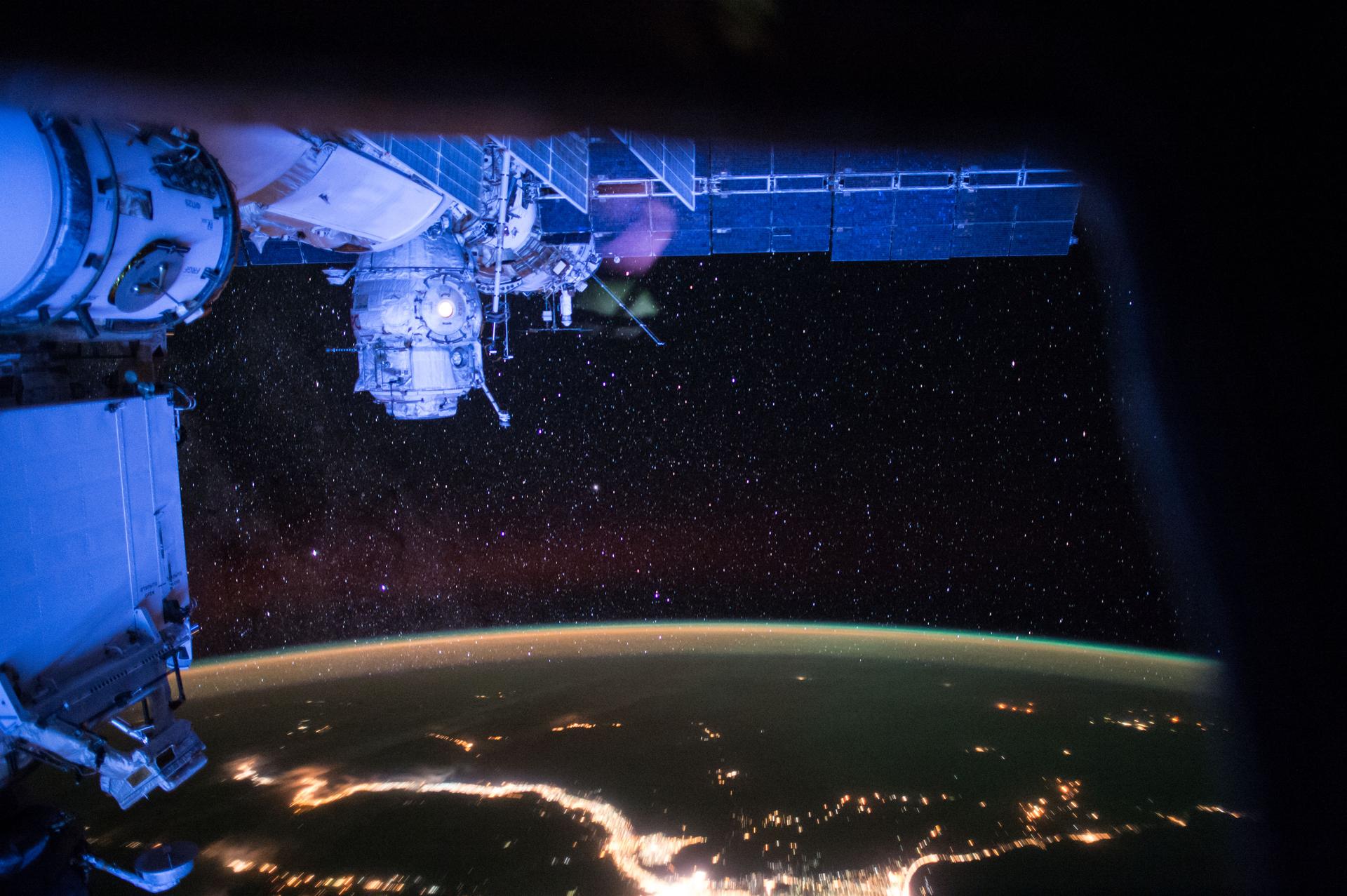Earth at night beneath the International Space Station, with city lights, stars, and aurora visible